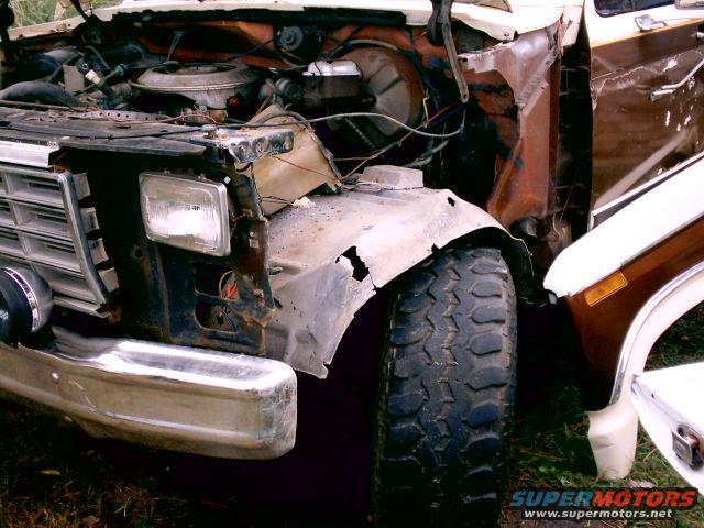 Front end of a damaged truck, showing a bent fender, exposed engine components, and a chrome bumper