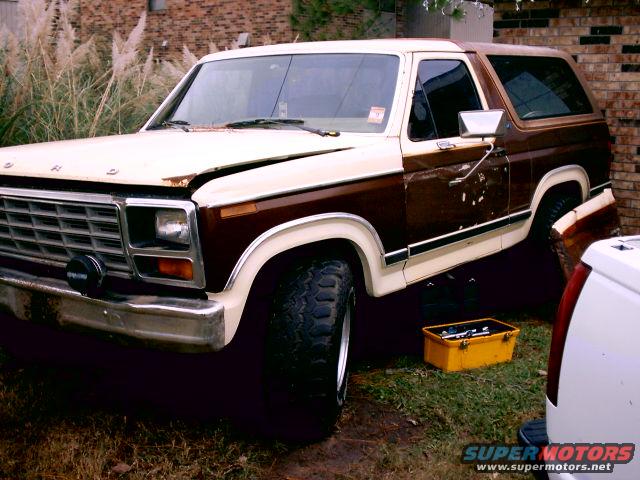 Two-tone brown and white Ford Bronco, front quarter view. Hood is open, showing some rust