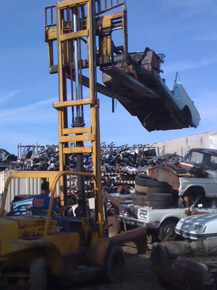 forklift lifting a car body over a junkyard with piles of scrap metal and other vehicles