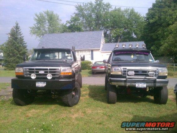 Two lifted 1994 Ford F-250 trucks parked in front of a house. The truck on the left has a large aftermarket bumper