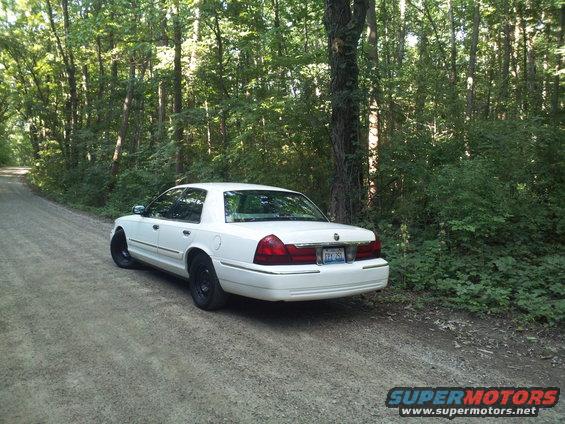 White sedan parked on a dirt road in a wooded area. License plate is visible