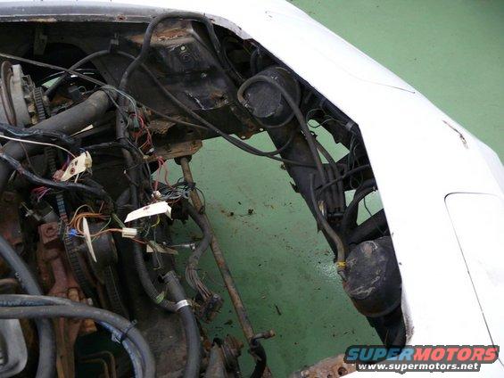 Car engine bay with exposed wiring and hoses, part of a white vehicle visible
