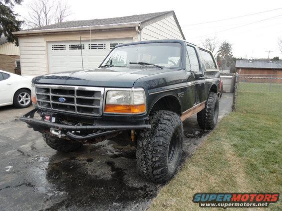 Dark green Ford Bronco with large off-road tires and a winch on the front bumper, parked in a driveway next to a garage