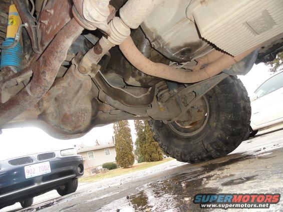 underneath a vehicle showing suspension, exhaust, and a large tire; another car is parked nearby