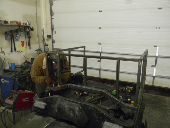 Welder in brown jacket and mask works on a metal frame attached to a black vehicle bed