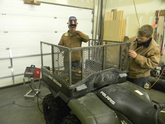 Polaris ATV with a custom metal cargo rack being welded on. Two men are working on the rack