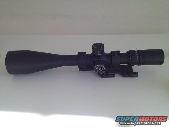 Black rifle scope with magnification adjustment turrets and a circular logo on a white surface