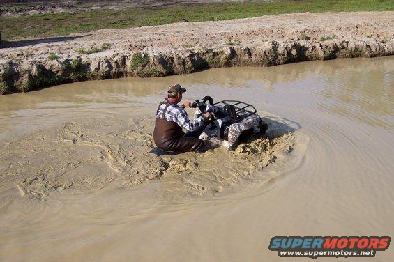 Guy in waders riding a camo ATV through a muddy water hole