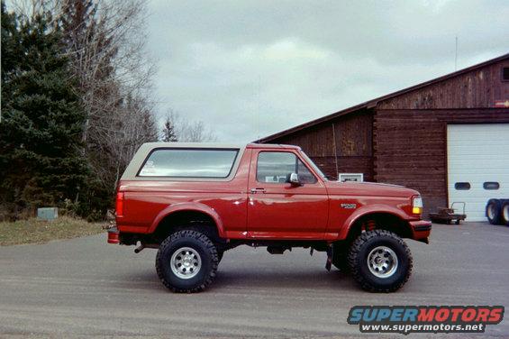 Red Ford Bronco with a tan top, lifted with large tires, parked on asphalt in front of a wooden building