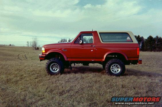 Red Ford Bronco, lifted with aftermarket wheels, parked in a grassy field