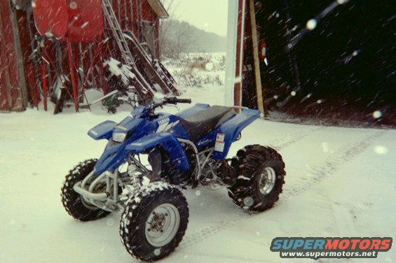 Blue Yamaha ATV parked in snow near a red barn with skis stacked