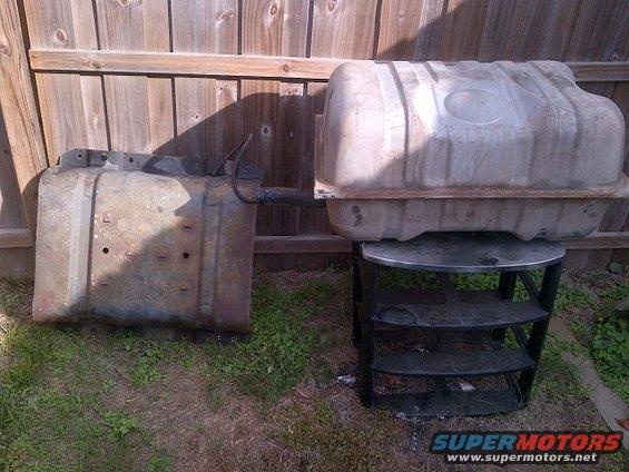Two old, rusty fuel tanks sit on the grass in front of a wooden fence. One is on the ground, the other is on a black shelf unit