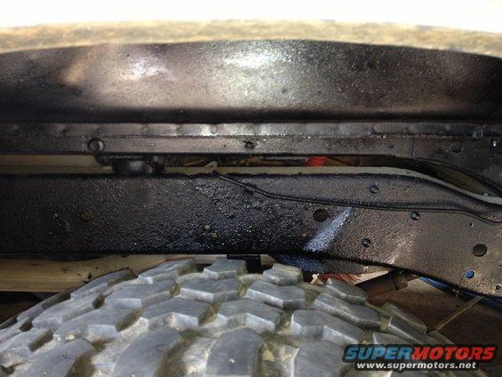 Close-up of vehicle frame and tire, showing black undercoating and part of a red shock absorber