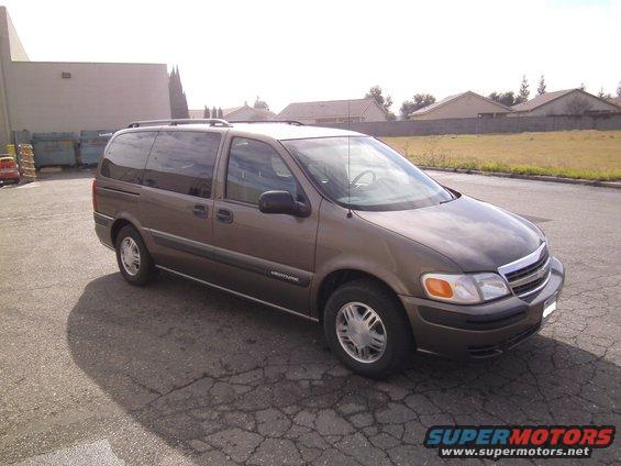 Brown minivan parked on cracked asphalt, with a building and some houses in the background