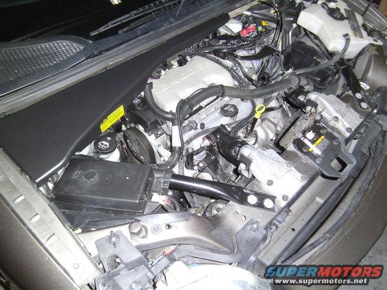 Engine bay with V6 engine, hoses, and electrical components visible. Some yellow warning labels are present