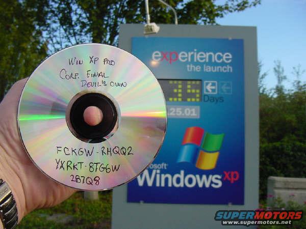 Hand holding CD with 'Win XP Pro Corp. Final Devil's Own' written on it, in front of a sign for Windows XP