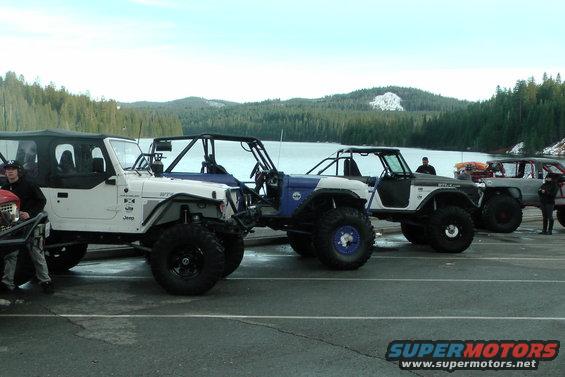 Several modified Jeeps and a truck parked by a lake with trees and snow-capped mountains in the background