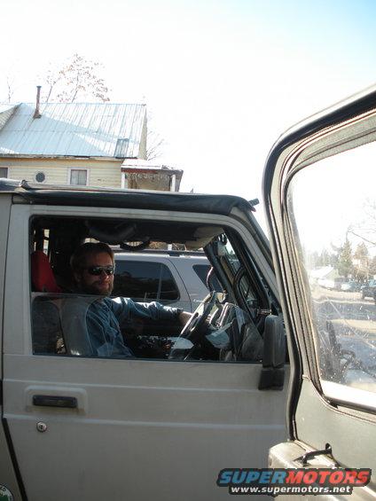 Man wearing sunglasses and a blue shirt sits in the driver's seat of a silver SUV. Another SUV is visible behind it