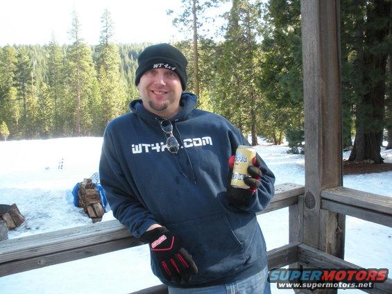 Man in beanie and hoodie holding a beer can, standing on a wooden deck with snow and trees in background