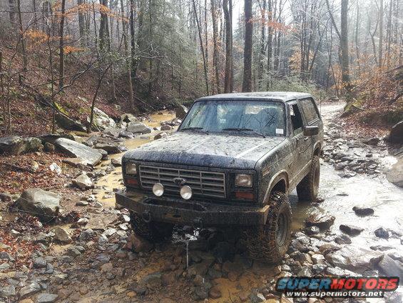 Dark blue Ford Bronco driving through a rocky stream bed in a forest