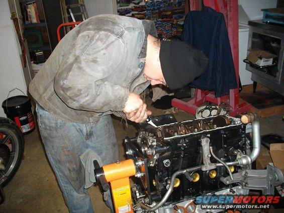 Engine block on an orange stand, being worked on by a person in jeans and a jacket. A Supermotors logo is visible