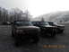 1986 Ford Bronco - Three Ford Broncos are parked in a gravel lot with trees and a misty hillside in the background.
