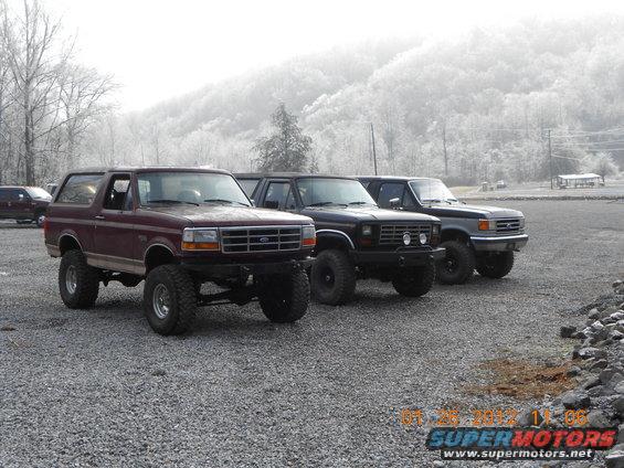 Maroon Bronco and two dark trucks parked on gravel lot with misty hills in background