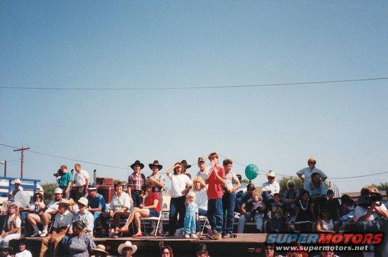 Crowd gathered on a wooden platform under a clear blue sky, some people wearing cowboy hats