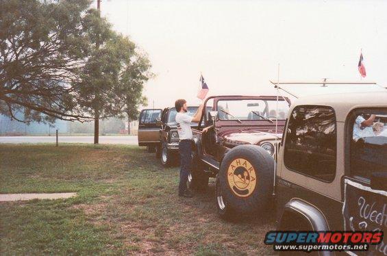 Line of SUVs and a Jeep parked on grass, with a man holding a flag