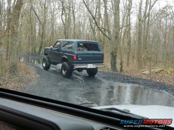 Dark green Ford Bronco driving on a muddy trail through woods. Lifted suspension and large tires