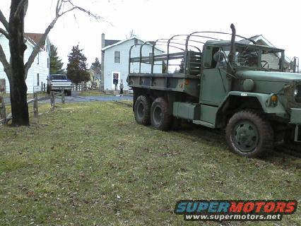Green military truck with a canvas top, parked on grass. Another truck is visible in the background