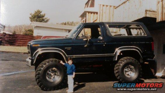 Side view of a dark blue 1995 Ford Bronco with large tires and a child standing beside it