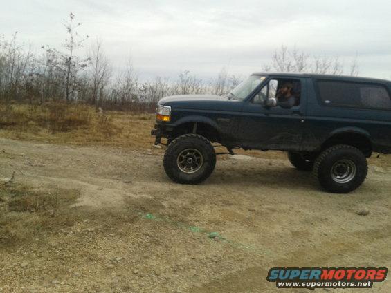 Dark green Ford Bronco with lifted suspension driving on a dirt road. Driver visible inside