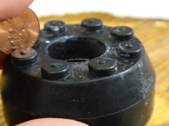 Black rubber part with six protruding studs, a penny is held next to it for size comparison