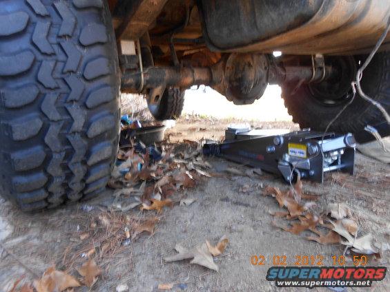 Undercarriage of a vehicle, showing a large tire, leaf springs, differential, and a hydraulic jack on the ground