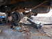 1986 Ford Bronco - The rear axle and suspension components of a vehicle are visible, supported by jack stands and a hydraulic jack, with leaves scattered on t…