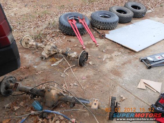 Two vehicle axles and four tires are laid out on concrete with a floor jack and some red tools