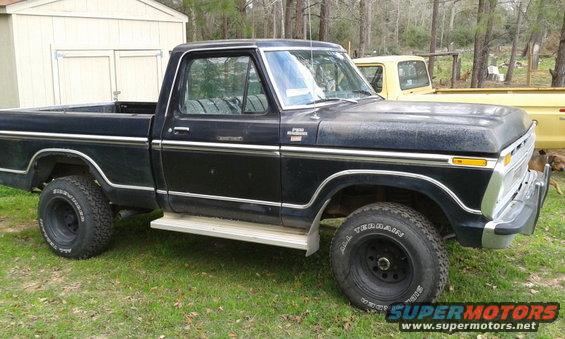 Black 1977 Ford F-150 pickup truck parked on grass next to a shed, with a yellow truck behind it