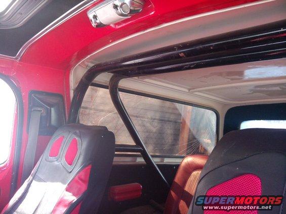 Interior view of a vehicle with red trim, black roll cage, and racing seats. A watermark reads 'SUPERMOTORS'