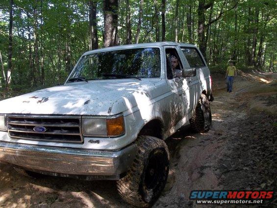 1990 Ford Bronco driving on a muddy forest trail with a person walking in the background