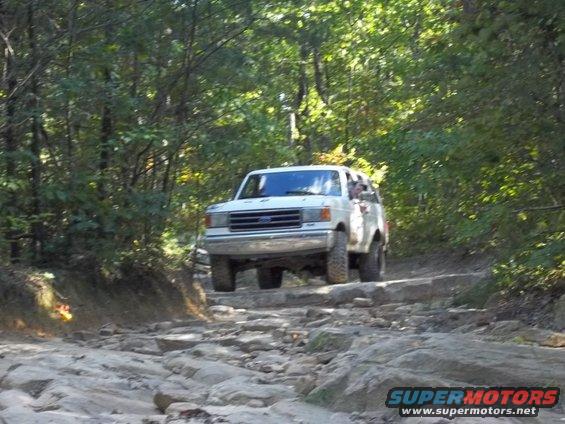 White Ford Bronco driving over rocks on a wooded trail. The vehicle appears to be a 1990 model