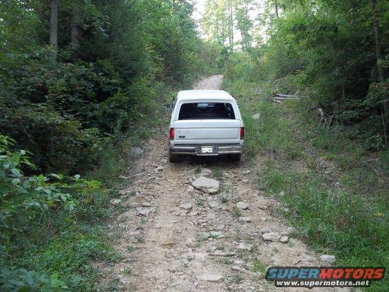 White 1990 Ford Bronco driving up a rocky, wooded dirt road