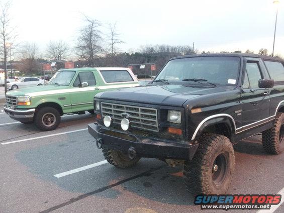Black Ford Bronco with aftermarket bumper and large tires, parked next to a green Bronco