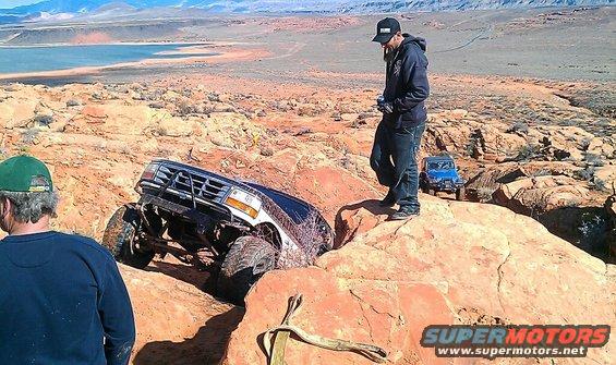 Off-road vehicle stuck on rocks, another vehicle behind it. Two people watching