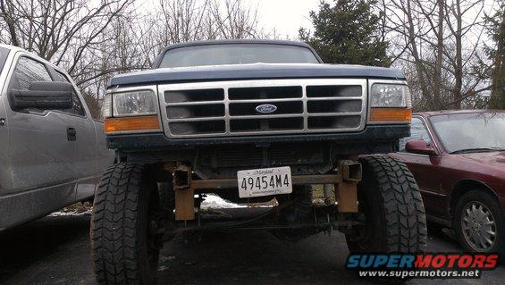 Front view of a lifted dark blue Ford truck with a chrome grille and a Maryland license plate. Another truck is to the left and a sedan to the right