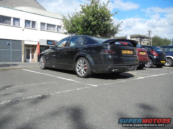 Black sedan with spoiler and dual exhaust pipes parked in a lot. Other cars are visible behind it