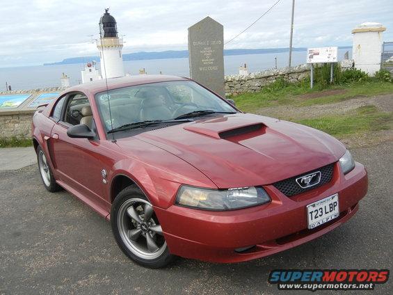 Red Ford Mustang with hood scoop parked near a lighthouse and a stone monument