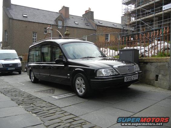 Black hearse with license plate OE02 ZTY parked on cobblestone street, white van in background