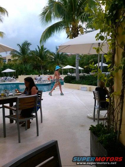 People by a pool at a resort, with palm trees and umbrellas. A woman walks past a woman sitting at a table