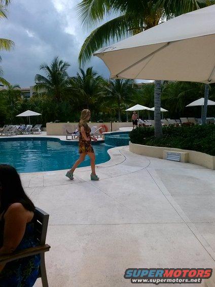 Woman in a patterned dress walks by a resort pool with palm trees and lounge chairs under a cloudy sky